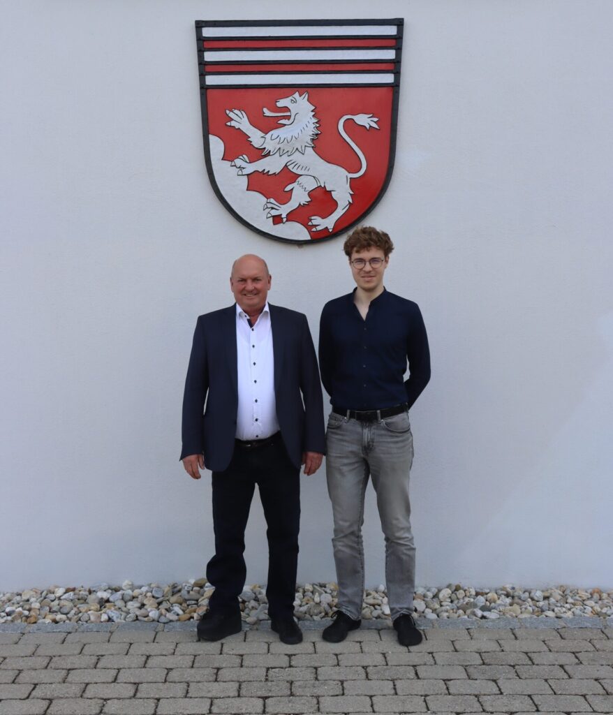 Bürgermeister Josef Moll und Klimaschutzmanager Adrian Heider stehen vor dem Wappen der Gemeinde Leiblfing.